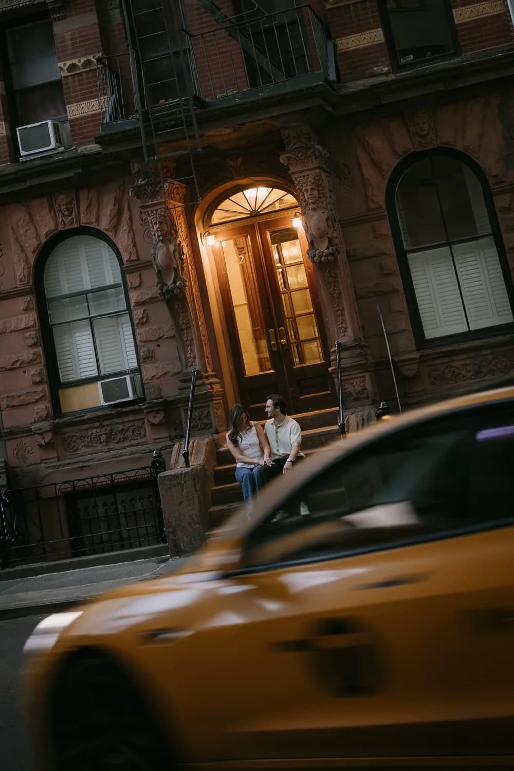 Rachel and Hugh sitting on a brownstone stoop as a yellow cab passes